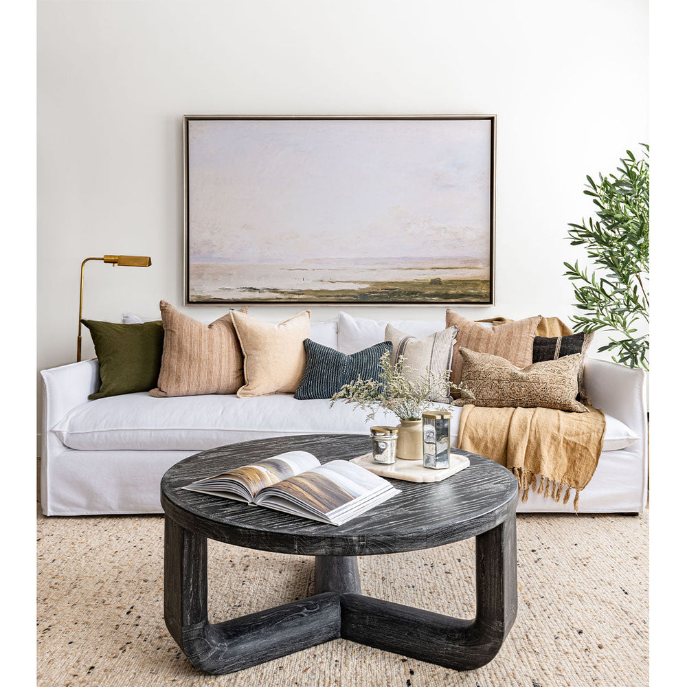 Lifestyle image of a living room with customizable white linen upholstery and textural, bohemian throw pillows with dark, round coffee table, painting, rug and olive tree.