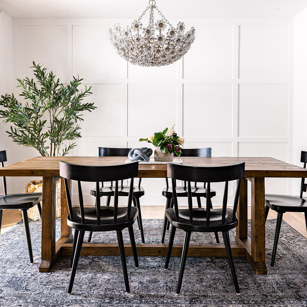 Formal dining room with wall panelling, crystal chandelier, olive tree, and black dining chairs around a wooden table.