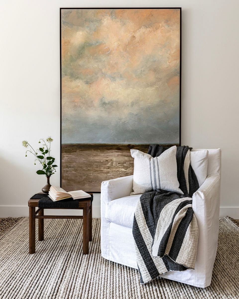 Large vertical artwork with blue-grey and a touch of peachy clouds, and murky grey waters. Styled with a white swivel chair, a black bench, some greenery and accessories.