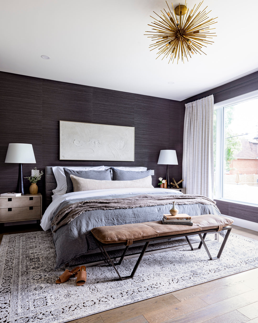 This master bedroom is spacious with dark tones coming through in the wallpaper, bedding, rug, and is enhanced with warm tones found in the light fixture, bench, and nightstands.