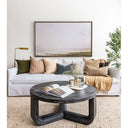 Lifestyle image of a living room with customizable white linen upholstery and textural, bohemian throw pillows with dark, round coffee table, painting, rug and olive tree.
