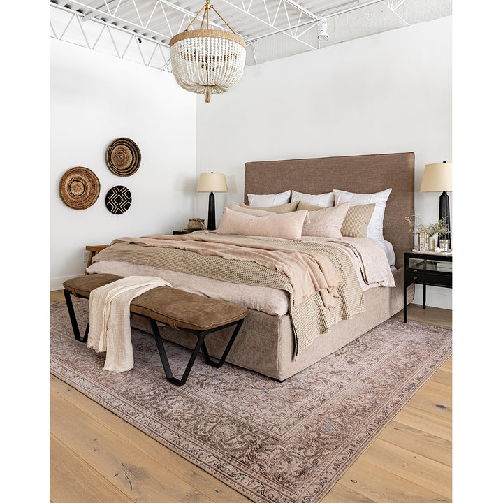 A lifestyle image of the Villa Bed in a king size. Styled with a leather bench, blush-toned antique rug, beaded chandelier, and blush pillows. Custom upholstery in a brown natural tone.